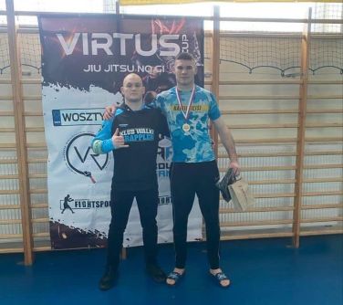 Hala sportowa. Dwóch mężczyzn pozuje do zdjęcia na tle drabinek na hali sportowej. Trzymają się za ramiona