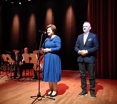 Scena. Kobieta w niebieskiej sukience przemawia do stojącego mikrofonu. Obok stoi mężczyzna. W tle na krzesłach orkiestra,...