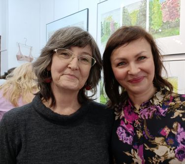 Pomieszczenie. Dwie uśmiechnięte kobiety pozują do zdjęcia. W tle na białej ścianie obrazy i osoby oglądające