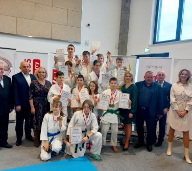 Hala sportowa. Na podium grupa dwunastu zawodników w kimonach. Trzymają dyplomy w dłoniach. Po lewej stoi troje dorosłych,...