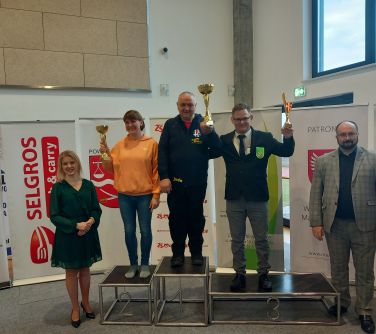 Hala sportowa. Na podium kobieta i dwóch mężczyzn trzymają puchary. Po lewej obok podium stoi kobieta. Po prawej mężczyzna