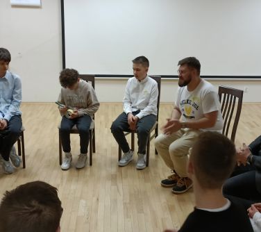 Sala konferencyjna. Naokoło na krzesłach siedzą nastoaltkowie i mężczyzna który prowadzi warsztaty. W tle biała duża tablica