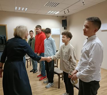 Sala konferencyjna. Pięcioro nastolatków stoi przed krzesłami, uśmiechnięta kobieta podaje chłopcu rękę