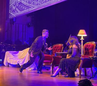 Scena. Na niej sofa, dwa fotele pomiędzy którymi stoją dwa stare gramofony, zapalona stojąca lampa. Kobieta w czarnej sukni...