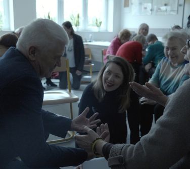 Sala lekcyjna. Na krzesłach siedzą osoby, rozmawiają gestykulując.