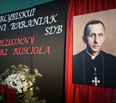 W pomieszczeniu. Na ścianie na czarnym tle tytuł Arcybiskup Antoni Baraniak SDB Niezłomy Pasterz Kościoła. Przed kwiaty...
