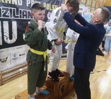 Hala sportowa. Na podium dzieci. Mężczyzna w granatowej marynarce zakłada medal dziecku na szyje za pierwsze miejsce