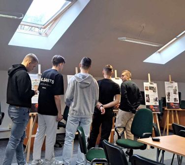 Pięciu nastolatków w pomieszczeniu ogląda wystawę ustawioną na drewnianych sztalugach. W suficie otwierane duże okna