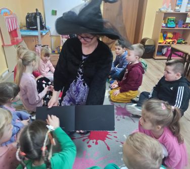 Sala lekcyjna. Dzieci siedzą na podłodze. Między nimi kobieta przebrana w czarny kapelusz z tiulem, pokazuje czarną kartkę