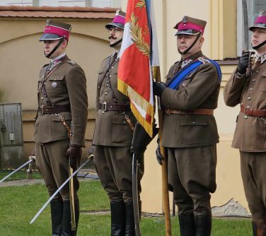 Na dworze. Czterech ułanów w mundurach ze sztandarem z szablami w dłoniach stoi na chodniku