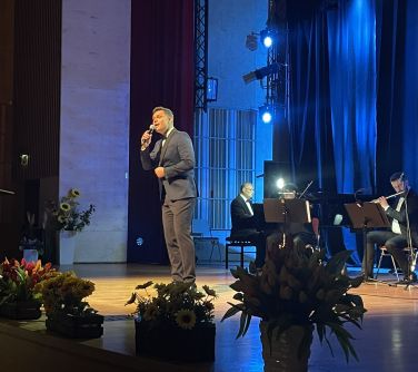 Pomieszczenie. Na scenie śpiewa mężczyzna. Za nim orkiestra. Przed nim na scenie kwiaty