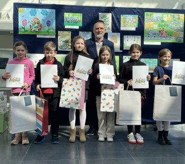 Pomieszczenie. Szcześcioro dzieci i mężczyzna pozują na tle granatowej płachty z pracami plastycznymi. Dzieci trzymają...