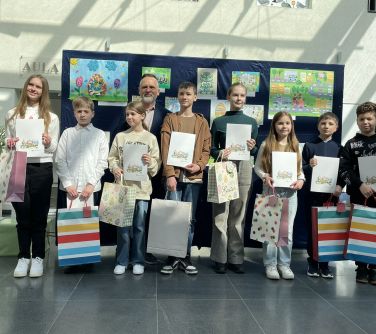 Pomieszczenie. Ośmioro dzieci i mężcyzzna pozują na tle grantowej płachty z pracami plastycznymi. Dzieci trzymają białe...