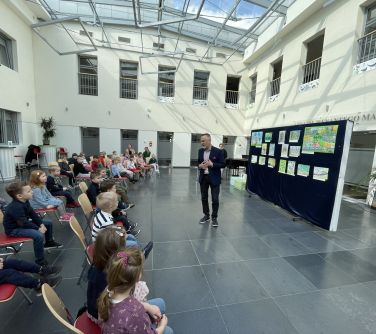 Pomieszczenie, przeszklony sufit. Dzieci siedzą na krzesłach. na przodzie przemawia mężcyzna. Za nim na niebieskiej...
