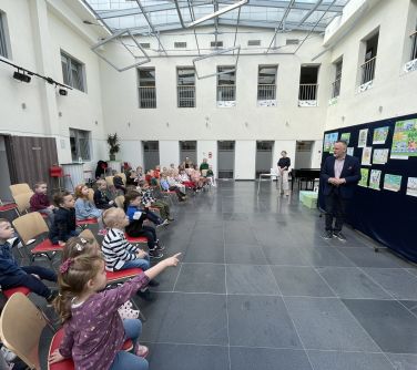 Pomieszczenie, przeszklony sufit. Dzieci siedzą na krzesłach. Na przodzie przy niebieskiej płachcie z pracami plastycznymi...