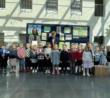 Pomieszczenie. Grupa dzieci z wychowawczynią i mężczyzną pozują na tle grantowej płachty z pracami plastycznymi. Po prawej...