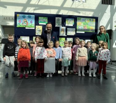 Pomieszczenie. Grupa dzieci z wychowawczynią i mężczyzną pozują na tle grantowej płachty z pracami plastycznymi. Po środku...