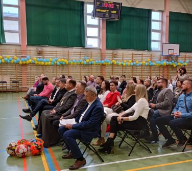 Hala sportowa. Na krzesłach siedzą osoby. W tle drabinki udekorowane balonami.