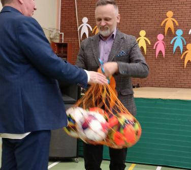 Hala sportowa. Mężczyzna z garniturze wręcza drugiemu mężczyźnie siatkę z pięcioma piłkami do piłki nożnej.