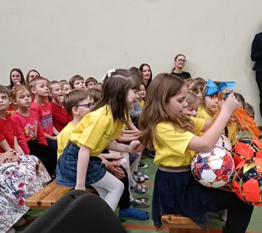 Hala sportowa. Na ławkach siedzą dzieci. Jedna z dziewczynek trzyma siatkę z piłkami do gry. Dziewczynka za nią zagląda jej...