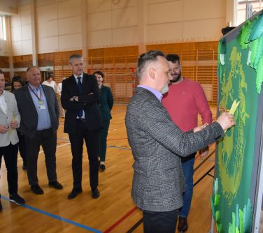 Hala sportowa. Mężczyzna w szarej marynarce stojąc bokiem przyczepia kartkę na zielonej tablicy. W tle stoją osoby