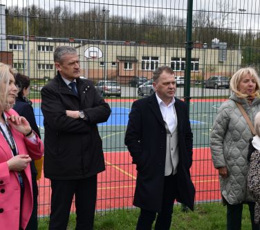 Na dworze. Pięć osób stoi w zapatrzeniu. W tle wysokie ogrodzenie boiska
