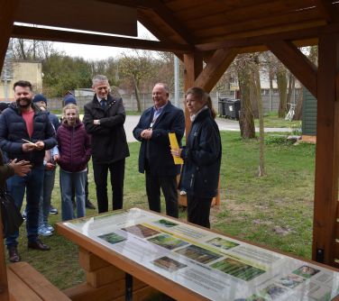 Na trawniku przed drewnianą wiatą stoi grupa uśmiechniętych osób. Patrzą na kolorową planszę leżacą na stole pod wiatą.