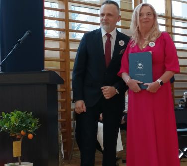 Hala sportowa. Mężczyzna w garniturze i kobieta w długiej różowej sukience pozują do zdjęcia. Kobieta ma granatową teczkę.