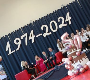 Hala sportowa. Na scenie na czerwonych sofach siedzi pięc osób. Po prawej na ławkach siedzą dzieci. Z przodu dekoracja w...