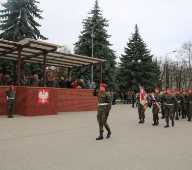 Na dworze. Na placu maszerują żołnierze z karabinami. Na przodzie prowadzący, salutujący żołnierz. Wszyscy patrzą w...
