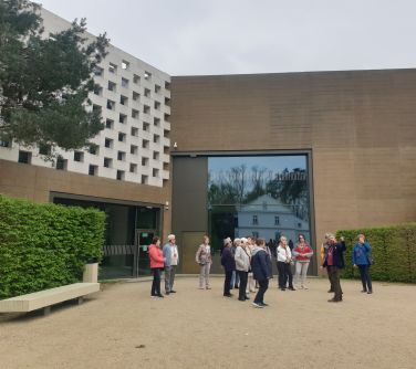 Grupa ludzi stoi na zewnątrz. W tle budynki muzeum.