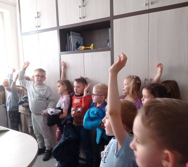 Pokój. Zgromadzone dzieci podnoszą ręcę do odpowiedzi. W tle szafy