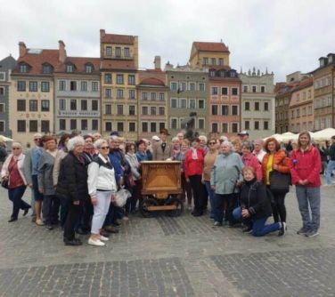 Na dworze, starówka. Grupa osób pozuje do zdjęcia. W środku katarynka i kataryniarz. W tle kolorowe kamienice.