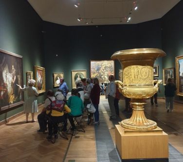 Wnętrze muzeum. Na środku na postumencie duży złoty kielich. W tle siedzą osoby i słuchają kobiety stojącej obok.