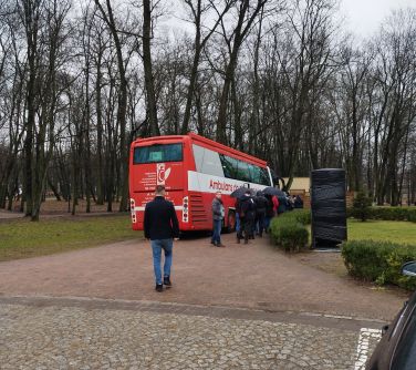 Na dworze. W alejce stoi biało-czerwony autobus. Przed autobusem kolejka osób do oddania krwi