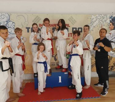 Hala sportowa. Ośmioro osób w kimonach w pozycji bojowej pozują do zdjęcia. Obok mężczyzna w czarnym dresie.