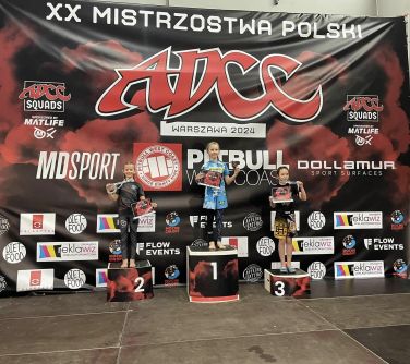 Hala sportowa.Na podium młode zawodniczki. W jednym ręku pokazuja medal zawieszony na szyi, w drugiej trzymają dyplomy