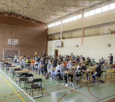 Hala sportowa. Widok z daleka na siedzącą na rozkładanych krzesłach młdzodzież. Grają w szachy.