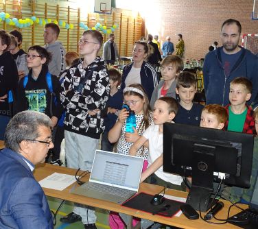 Grupa osób stoi przed stołami, na których stoją monitor i komputer.
