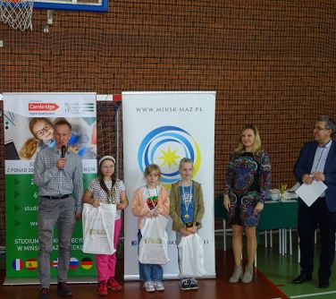 Troje dzieci w towarzystwie trójki dorosłych stoi w hali sportowej na tle rollupów. Jeden z mężczyzn mówi do mikrofonu.
