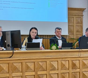 Sala. Przy długim stole siedzą cztery osoby. Kobieta w środku przemawia do mikrofonu. Czyta dokumenty. W tle ekran