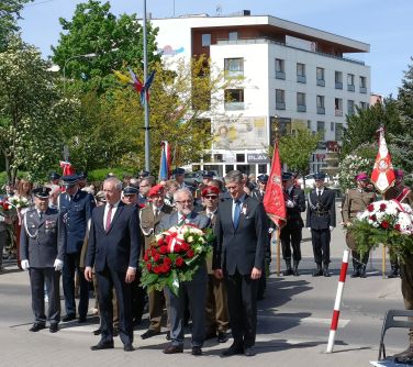 Dwór. Spora grupa ludzi w kolejce do złożenia kwiatów pod pomnikiem. W tle jasny blok i drzewa