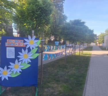 Dwór. Wzdłuż chodnika, na trawie, pomiędzy drzewami, na sznurkach rozwieszone kolorowe prace dzieci.