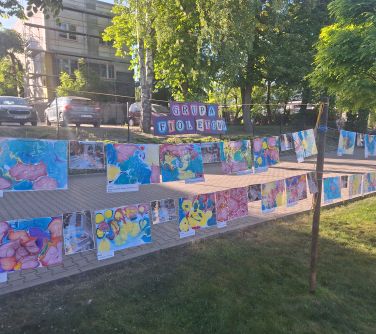 Dwór. Na trawie, pomiędzy młodymi drzewami na sznurkach wiszą kolorowe prace dzieci. W tle duże drzewa, auta i budynki