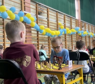 Sala gimnastyczna. Dwójka chłopcóe siedzi na przeciwko siebie przy stole. Na stole szachownica. Po lewej na drabinkach...
