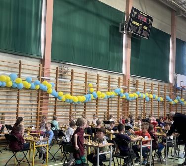 Sala gimnastyczna. Na stołach szachownice. Przy stołach dzieci rozgrywają parie. Po lewej żółto-niebieskie balony