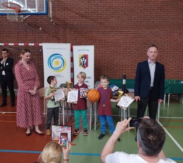 Sala gimnastyczna. Grupka dzieci otrzymała nagrody. Po prawej stoi mężczyzna w garniturze. Po lewejkobieta w sukience. W...