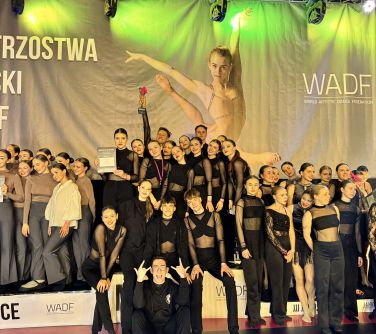 Pomieszczenie. Grupa uśmiechniętych tancerzy pozuje stojąc na podium na pierwszym miejscu. Z tyłu mężćzyzna podnosi puchar...