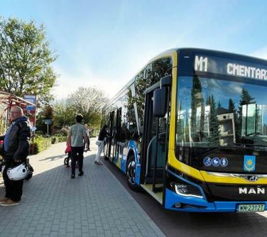 Na dworze. Po prawej autobus czarno-niebiesko-żółty. Po lewej osoby na przystanku