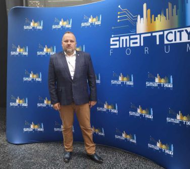 Pomieszczenie. Mężczyzna w ciemnej marynarce pozuje do zdjęcia na tle granatowej ścianki z tytułem smart city forum